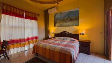 A hotel room with a bed, curtains, a painting, and a nightstand with a lamp.