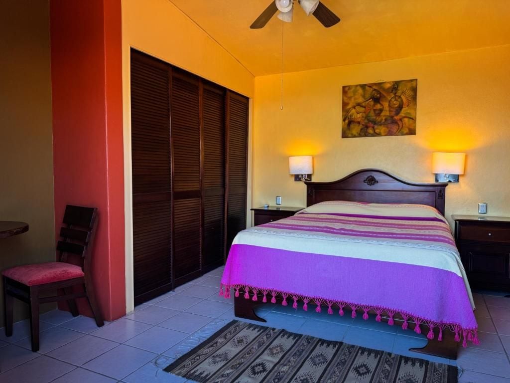 Habitación de hotel con cama, dos lámparas de noche, una silla, un ventilador de techo y un cuadro en la pared.