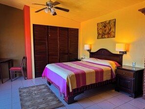 Habitación de hotel con cama, lámparas de noche, ventilador de techo, una silla, un tapete y un cuadro en la pared.