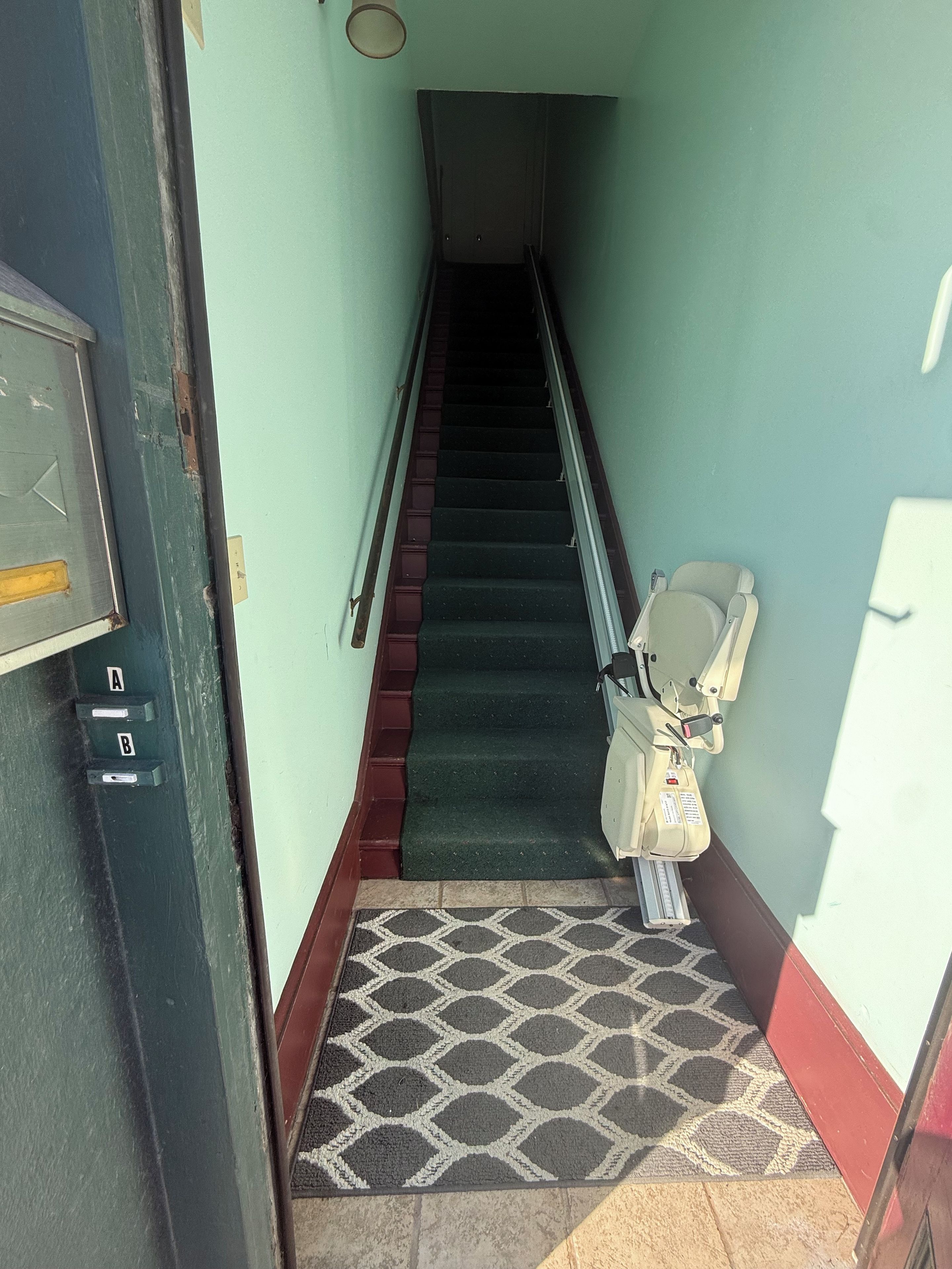 A staircase leading to a room with a door on the left, a handrail, and a patterned mat at the bottom.