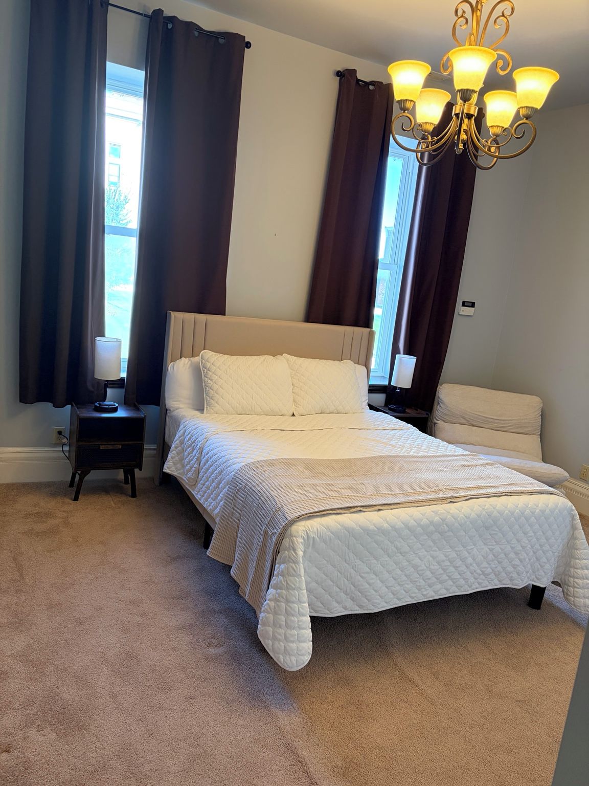 A bedroom with a bed, nightstands, a lamp, a chandelier, and curtains.