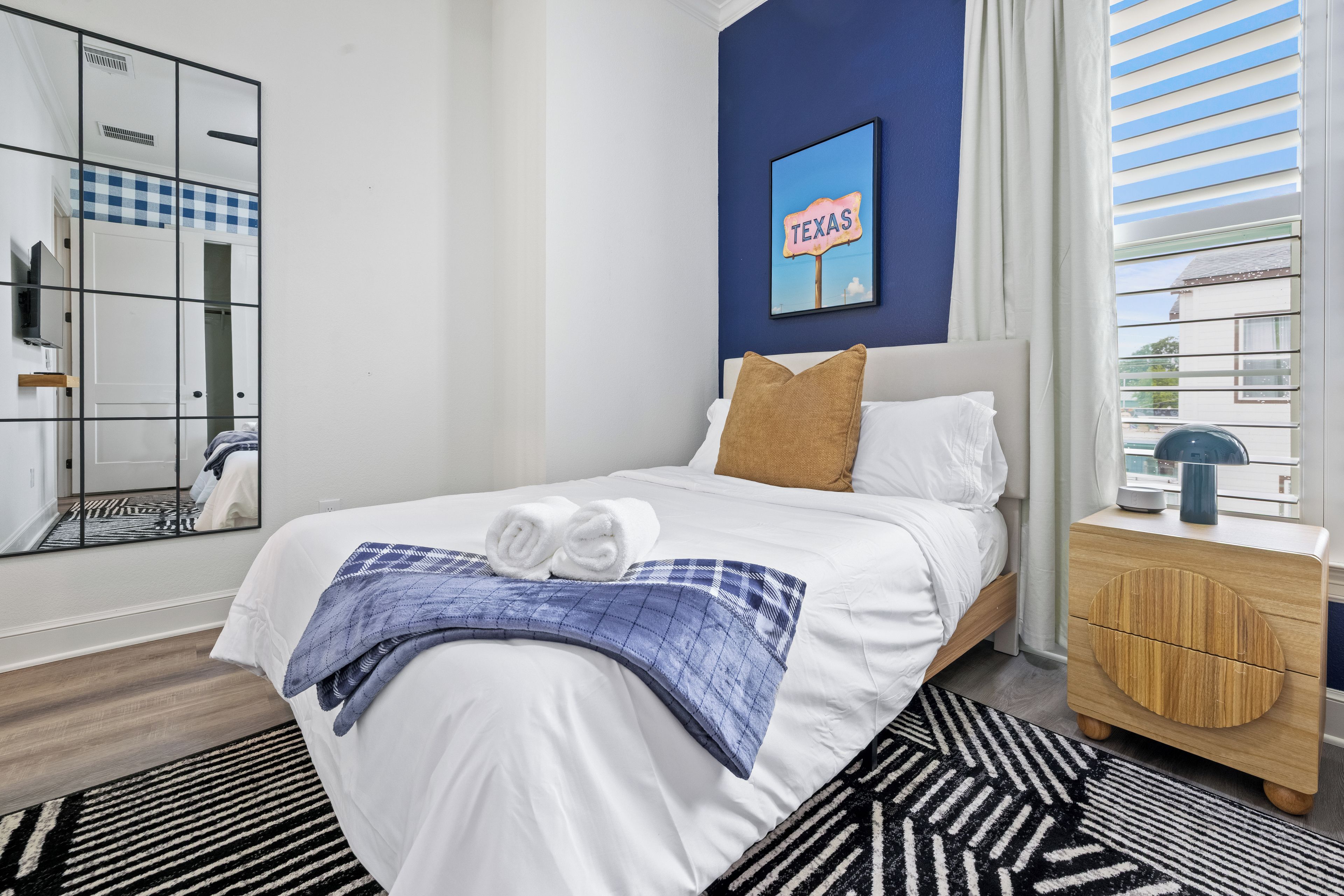 A hotel room with a bed, a nightstand, a mirror, a blue wall, and a window with blinds.