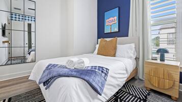 A hotel room with a bed, a nightstand, a mirror, a blue wall, and a window with blinds.