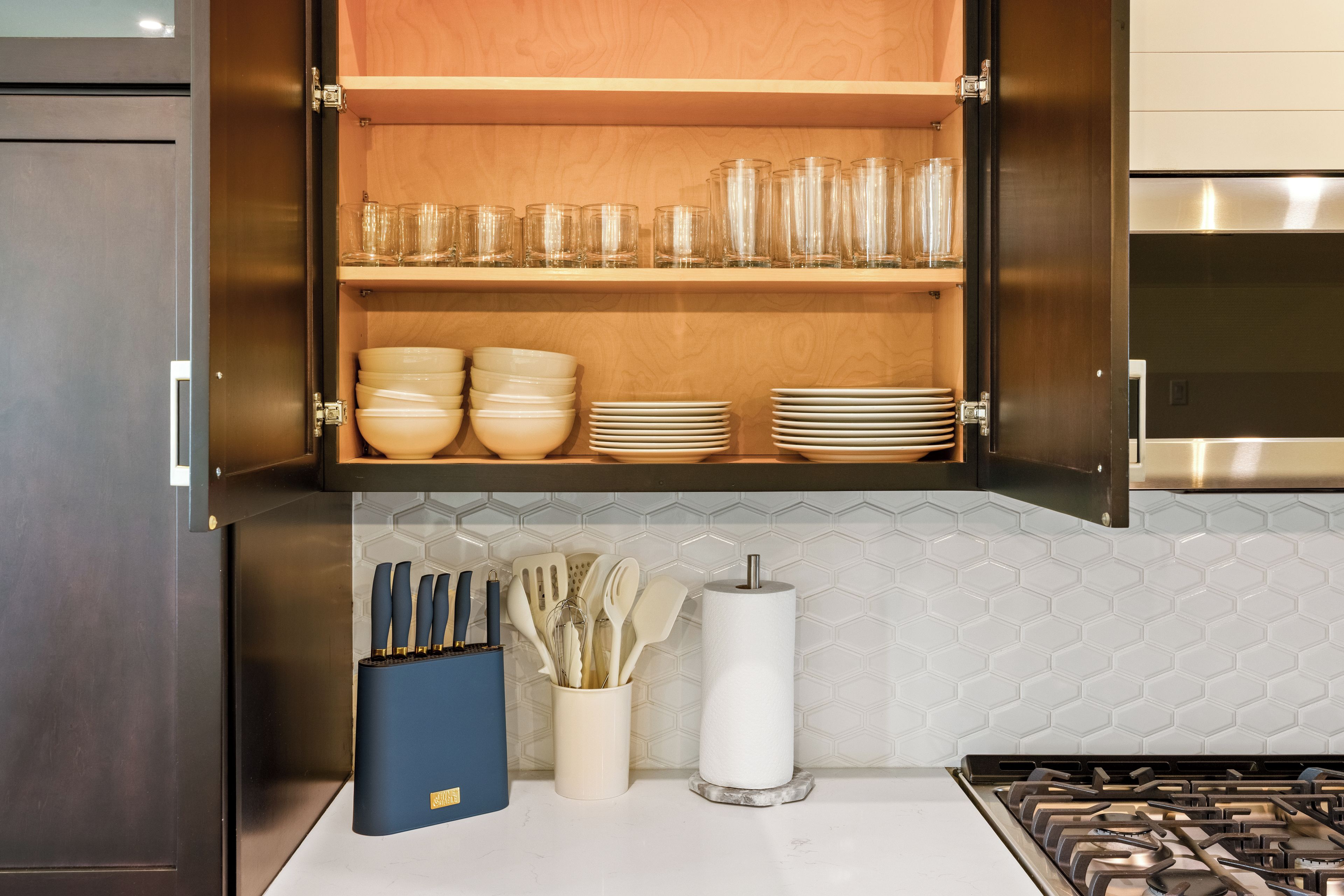 Una cocina con un armario de madera que contiene cristalería y cuencos. Debajo del armario, hay una encimera con un rollo de toallas de papel, un especiero y un caja de almacenamiento azul.