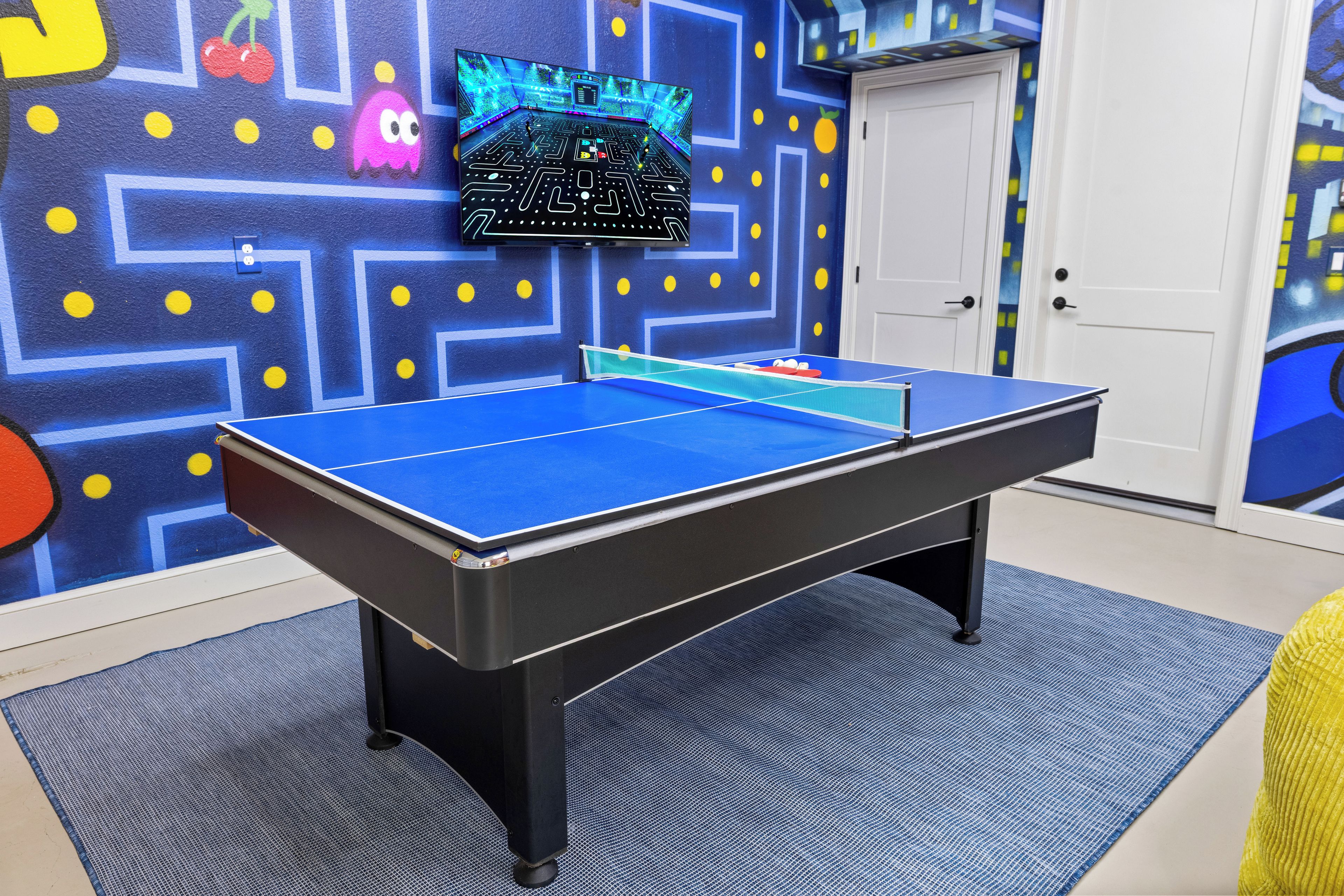 Una mesa de ping pong en una habitación con un mural azul y amarillo, una puerta y un mueble pequeño.