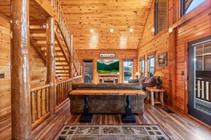 A log cabin interior with a wooden staircase, a sofa set, a television, and a window with a mountain view. - Cubs Dream - Mountain Views・Private Indoor Pool・Theater・Hot Tub・Arcade & More! (Sevierville)