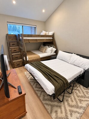 Quarto com beliche, escrivaninha com computador e controle remoto, e janela com vista para árvores.