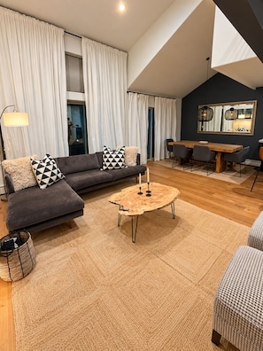 Een moderne woonkamer met een donkere bank, een ronde salontafel en een eethoek met een houten tafel en stoelen.
