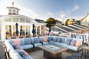 A deck with a built-in sofa and a glass-top coffee table, surrounded by wooden chairs and tables. - Inn at Good Harbor Beach (Gloucester)