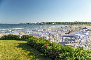A beach with striped umbrellas and lounge chairs, a grassy area with flowers, and a view of the sea and distant houses. - Inn at Good Harbor Beach (Gloucester)