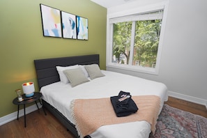 A neatly made bed with pillows and a blanket, a bedside table with a lamp, and a window with a view of trees.