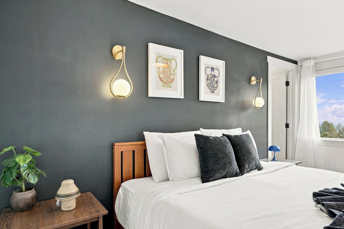 A bedroom with a bed, bedside tables, wall-mounted lights, framed artwork, a window with curtains, and a potted plant.