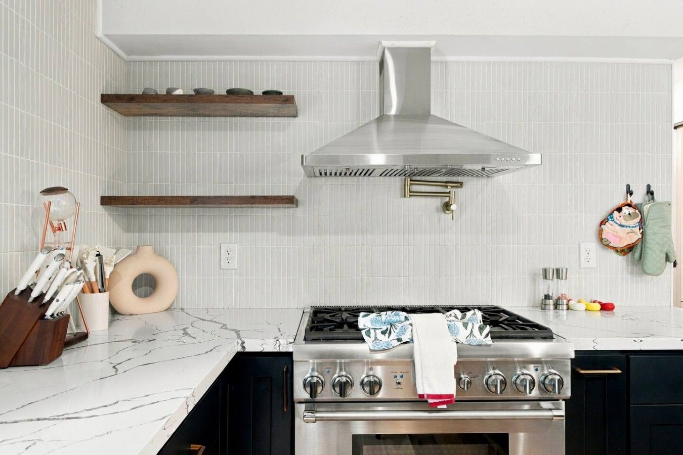 Une cuisine moderne dotée d’une cuisinière en acier inoxydable, d’étagères en bois et d’un dosseret de carrelage blanc.