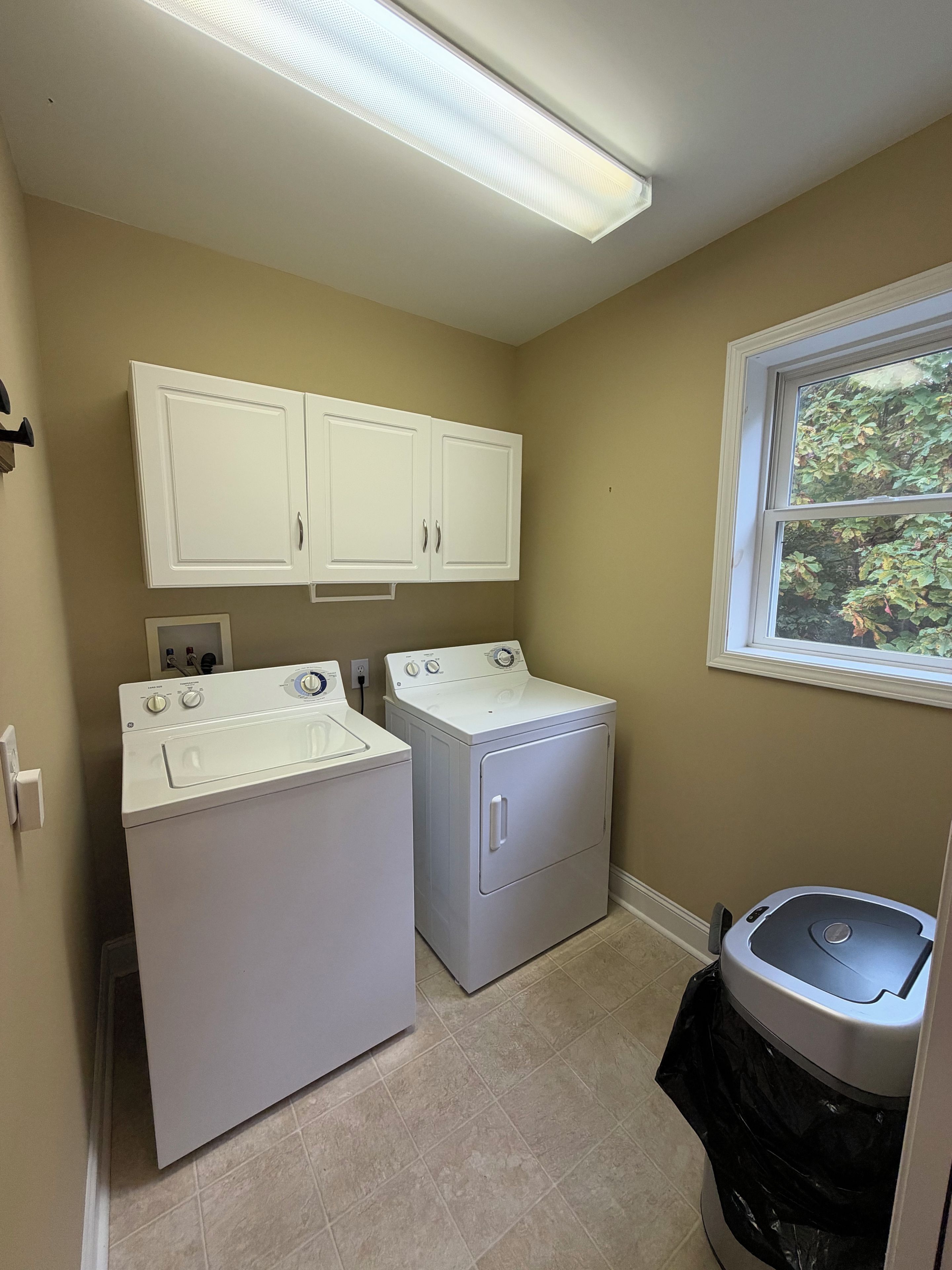 A laundry room with two washing machines, a dryer, and a trash can.