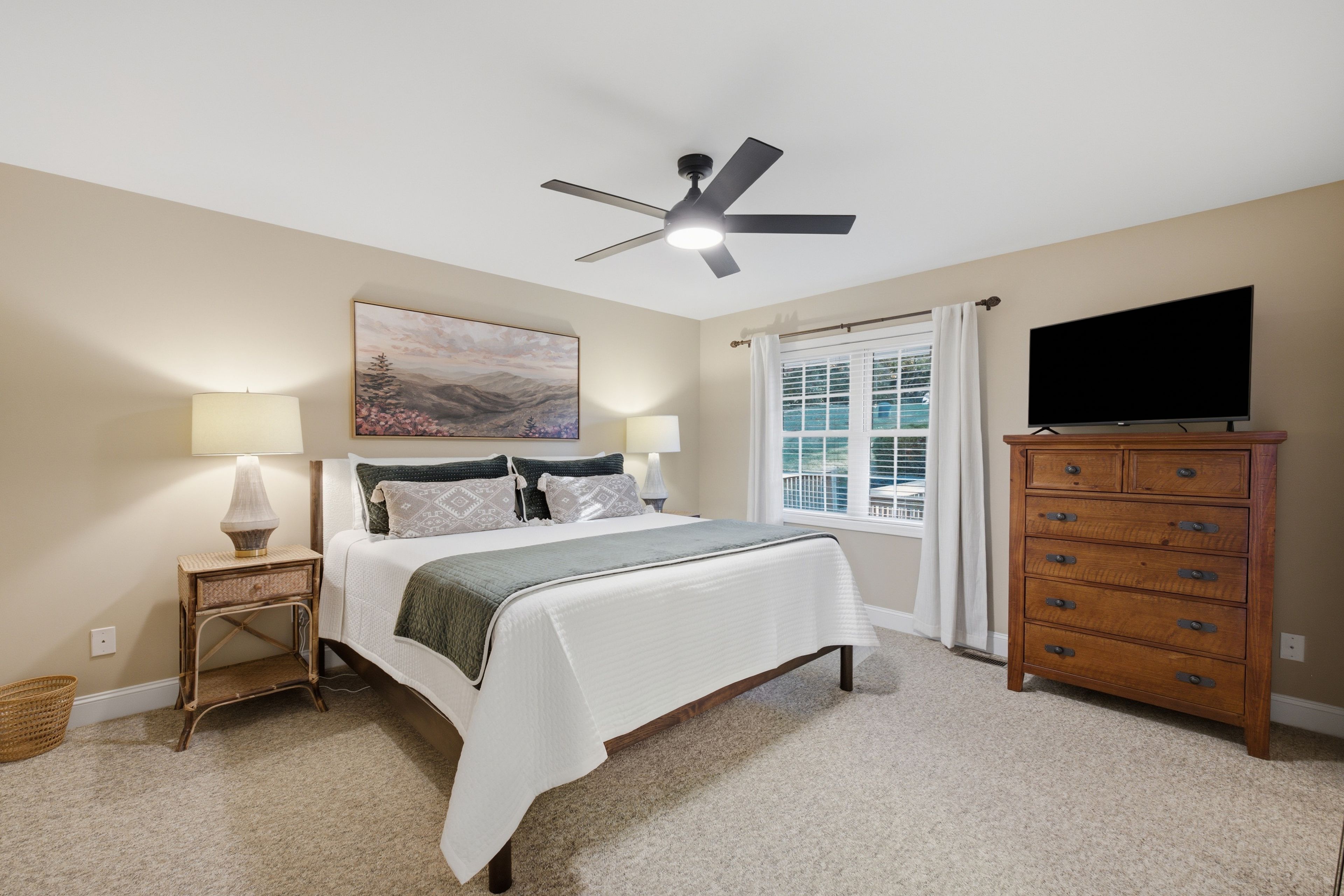Une chambre à coucher comprenant un lit, une table de chevet, une commode, un ventilateur de plafond, un tableau et une fenêtre avec des rideaux.