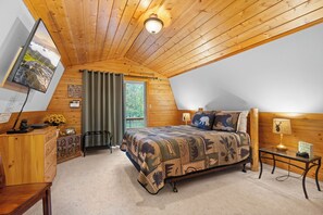Ein Schlafzimmer mit schrägem Dach, einem Bett mit gemustertem Quilz, einem Schreibtisch mit Computer und einem Fenster mit Vorhängen.
