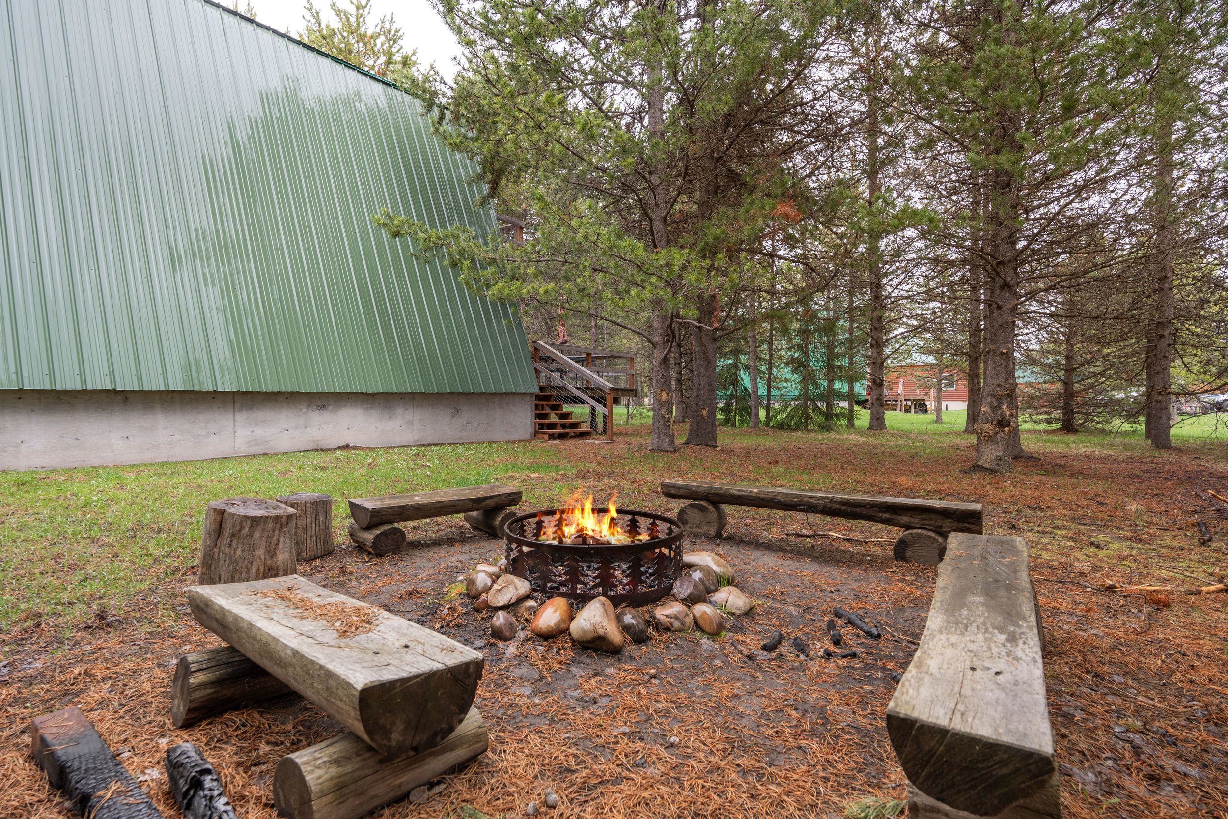Un foyer avec des bûches et des pierres, entouré de bancs en bois et d’une clôture en métal.