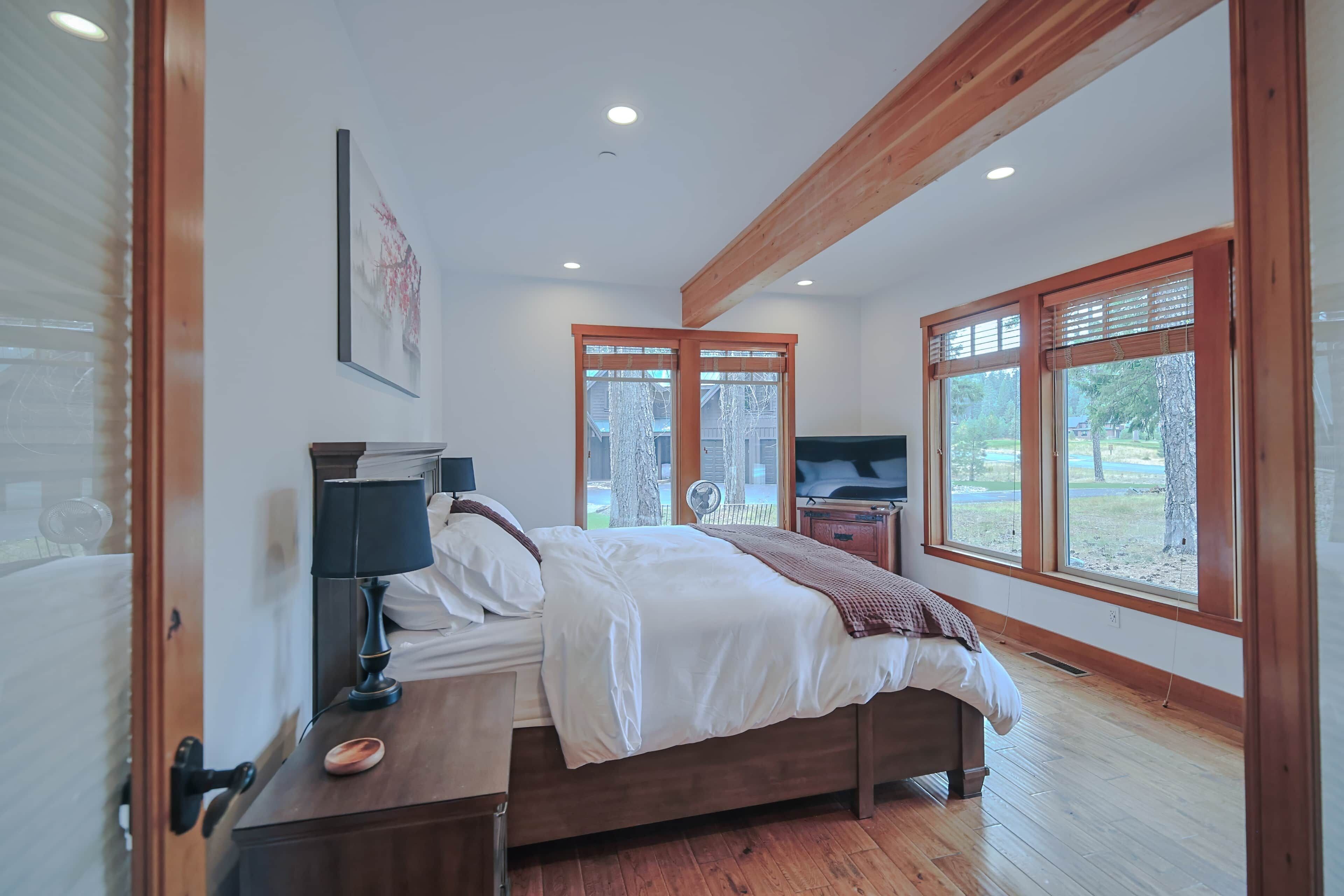 A bedroom with a bed, bedside table, lamp, and a large window with a view of the outdoors.