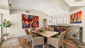 A dining area with a wooden table and wicker chairs, a colorful abstract painting, and a kitchen with white cabinets and a tiled backsplash. - Roadrunner Haven — warm and welcoming Monthly Rent (Scottsdale)