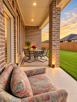 A covered patio with a brick wall, a round table with chairs, and a built-in bench with patterned cushions.