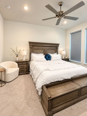 A bedroom with a large bed, a ceiling fan, bedside lamps, a chair, and a window.