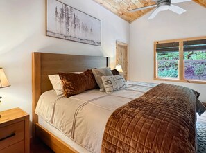 Ein Schlafzimmer mit einem hölzernen Kopfteil, einem Bett mit vielen Kissen, einem Nachttisch, einem Fenster mit Blick ins Grüne und einem Deckenventilator.