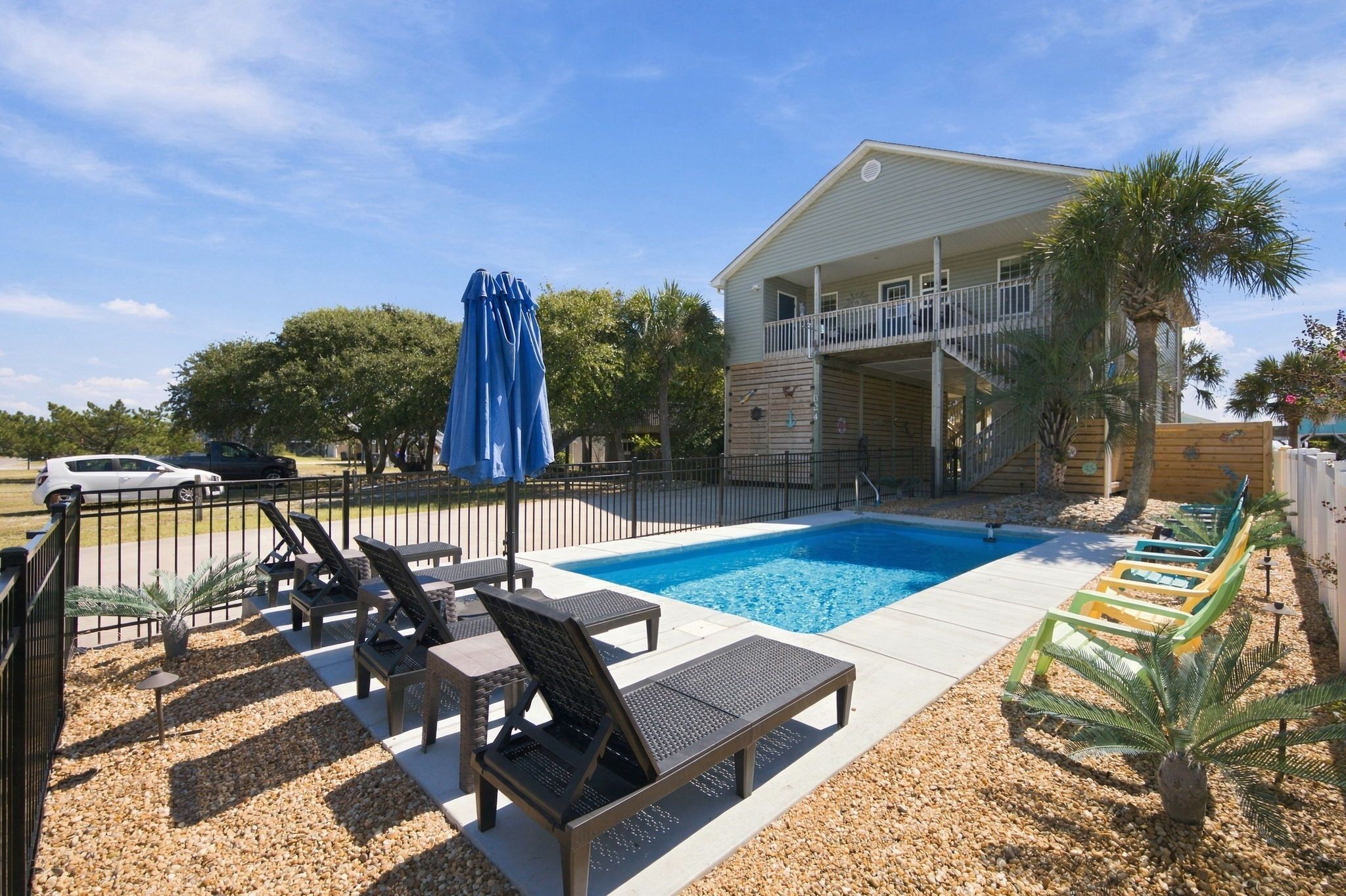 A swimming pool with lounge chairs and a covered umbrella, surrounded by a fenced area with palm trees.