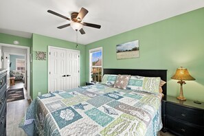 A bedroom with a bed, a ceiling fan, a nightstand with a lamp, and a door. - Sherry's Paradise: 4BR OKI Retreat, Heated Pool, Ocean Views, 2 Dogs Welcome! (Oak Island)