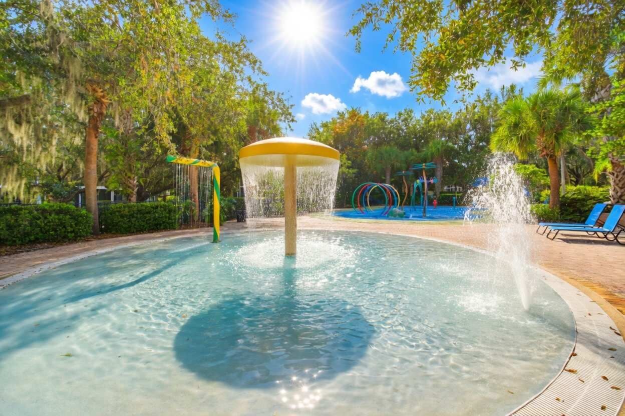 A circular water feature with a central fountain, surrounded by a paved area with lounge chairs and a playground in the background.