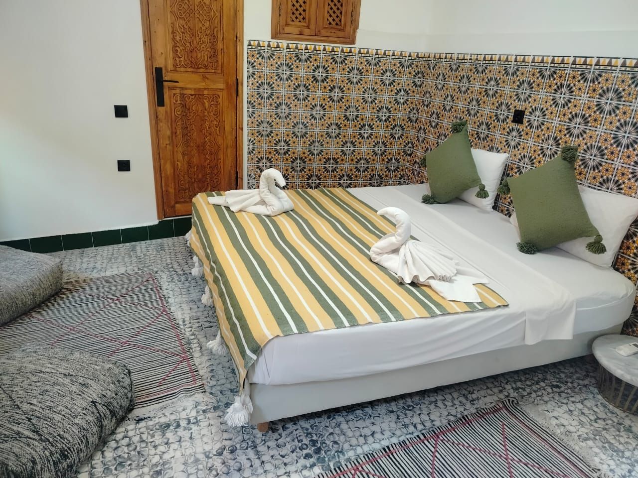 Une chambre d’hôtel avec un lit, une tête de lit ornée de motifs en carreaux, une porte en bois et une petite table ronde.