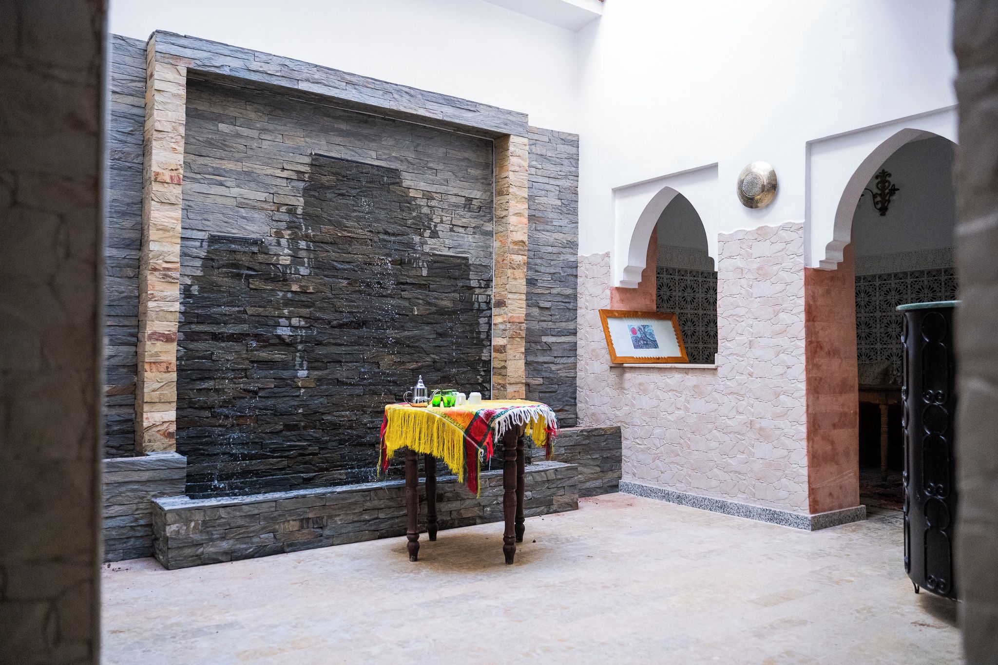 Une cour avec un mur en pierre, une table recouverte d’une nappe colorée et un tableau encadré accroché au mur.