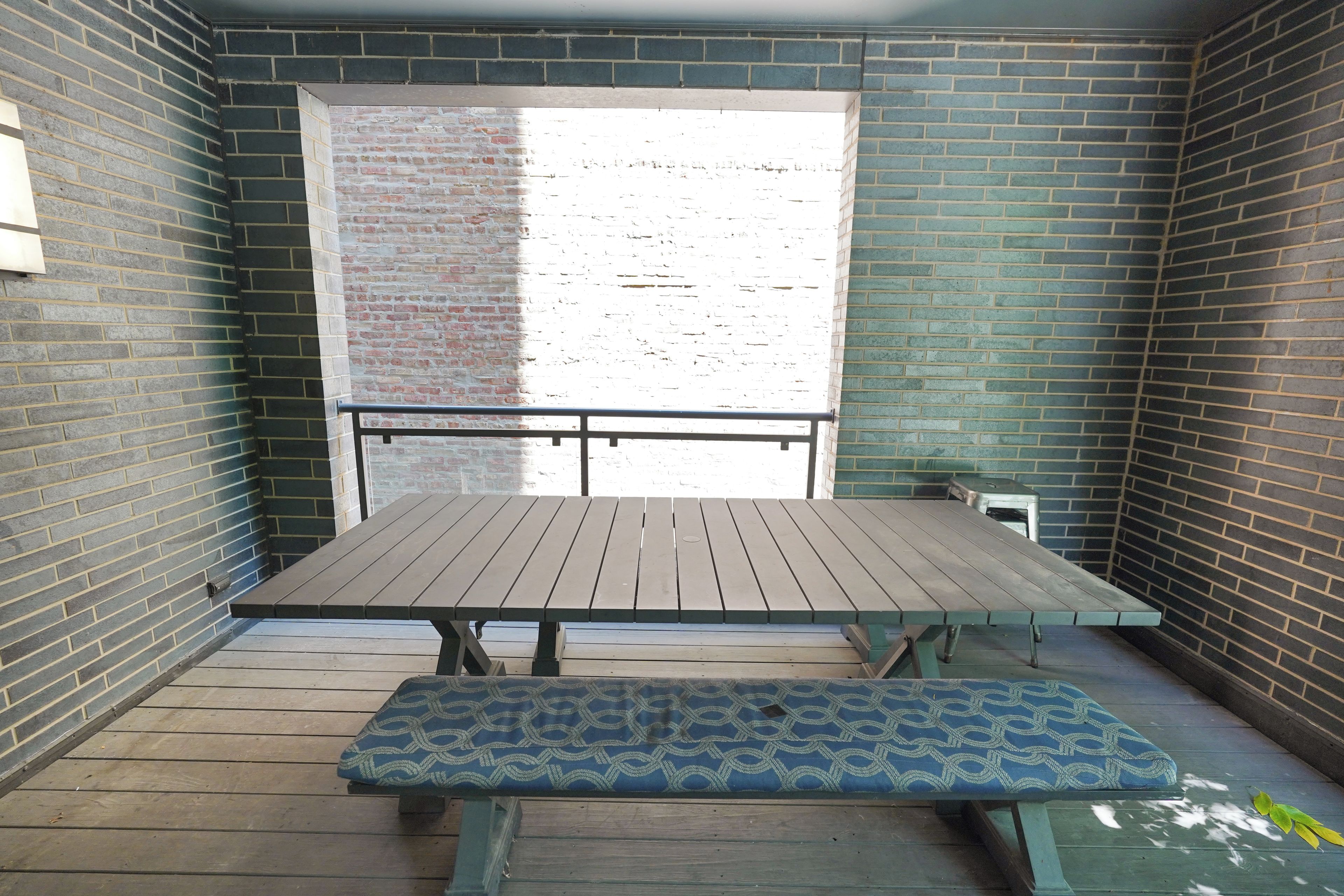 A balcony with a wooden table and bench, brick walls, and a view of a building.