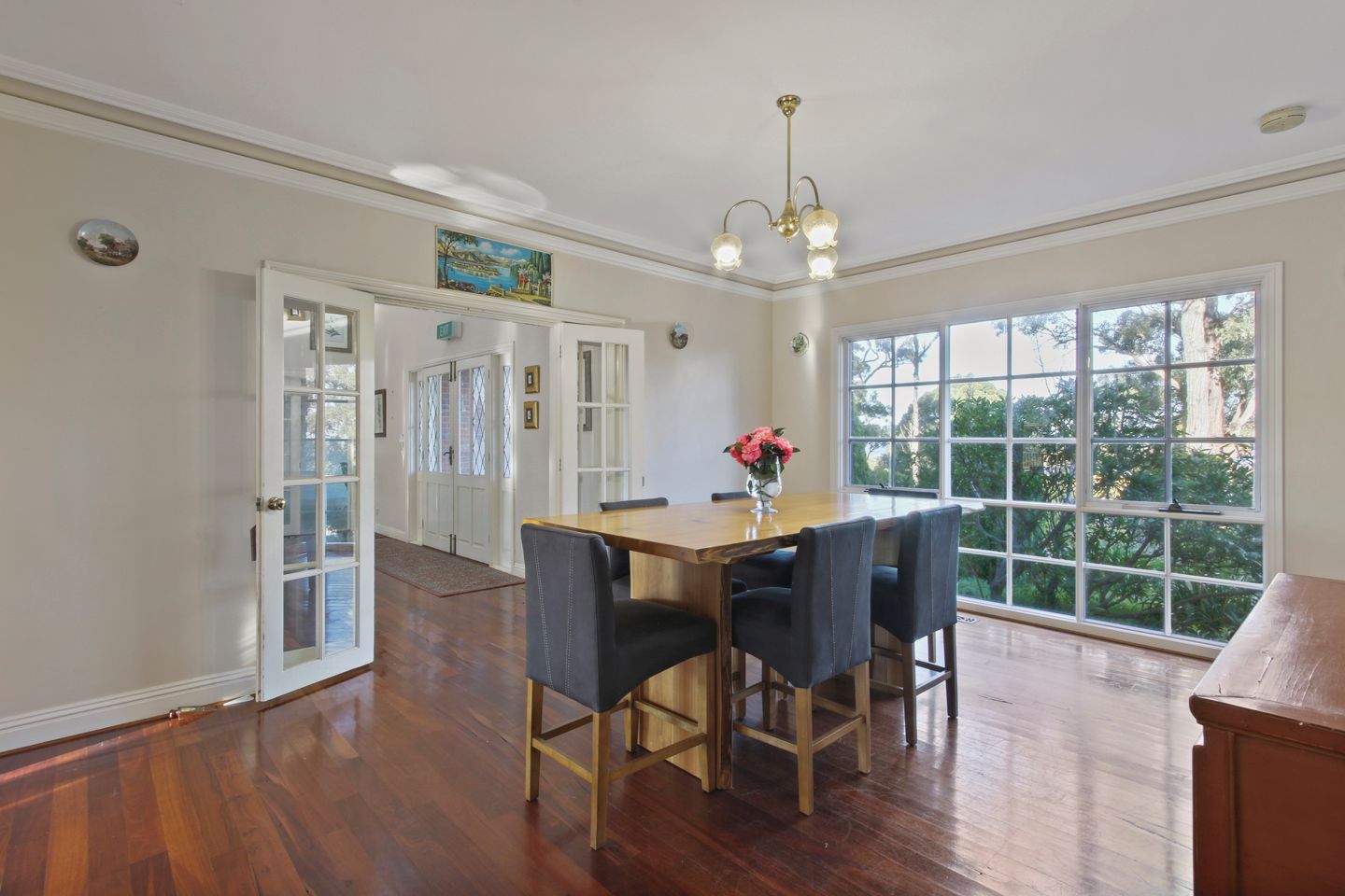A dining area with a wooden table and four chairs, a chandelier, and a view through large windows.