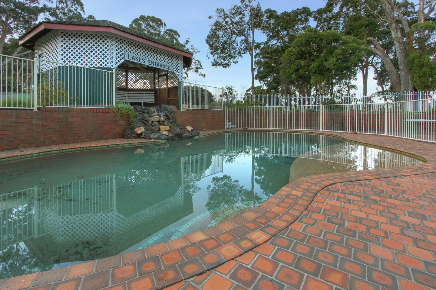 Una piscina con plataforma de ladrillo, rodeada por una valla blanca y un pabellón cubierto.