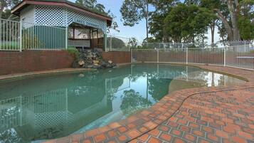 Una piscina con plataforma de ladrillo, rodeada por una valla blanca y un pabellón cubierto.