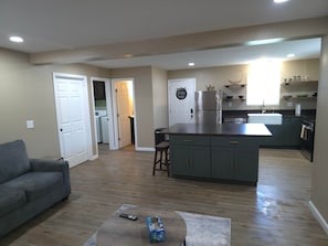 A modern kitchen with a central island, a dining area with a sofa, and a hallway leading to a laundry room. - Your Home Away From Home- Private Oasis (Wichita)