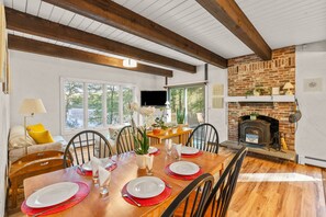 A dining area with a wooden table set for four, a brick fireplace, and a television in the background. - Mashpee Retreat 3BR 2BA Pond Access & Water Views (Mashpee)