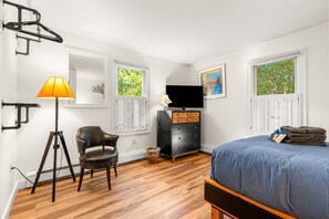 Quarto com cama, cadeira, escrivaninha, televisão e janelas com persianas.