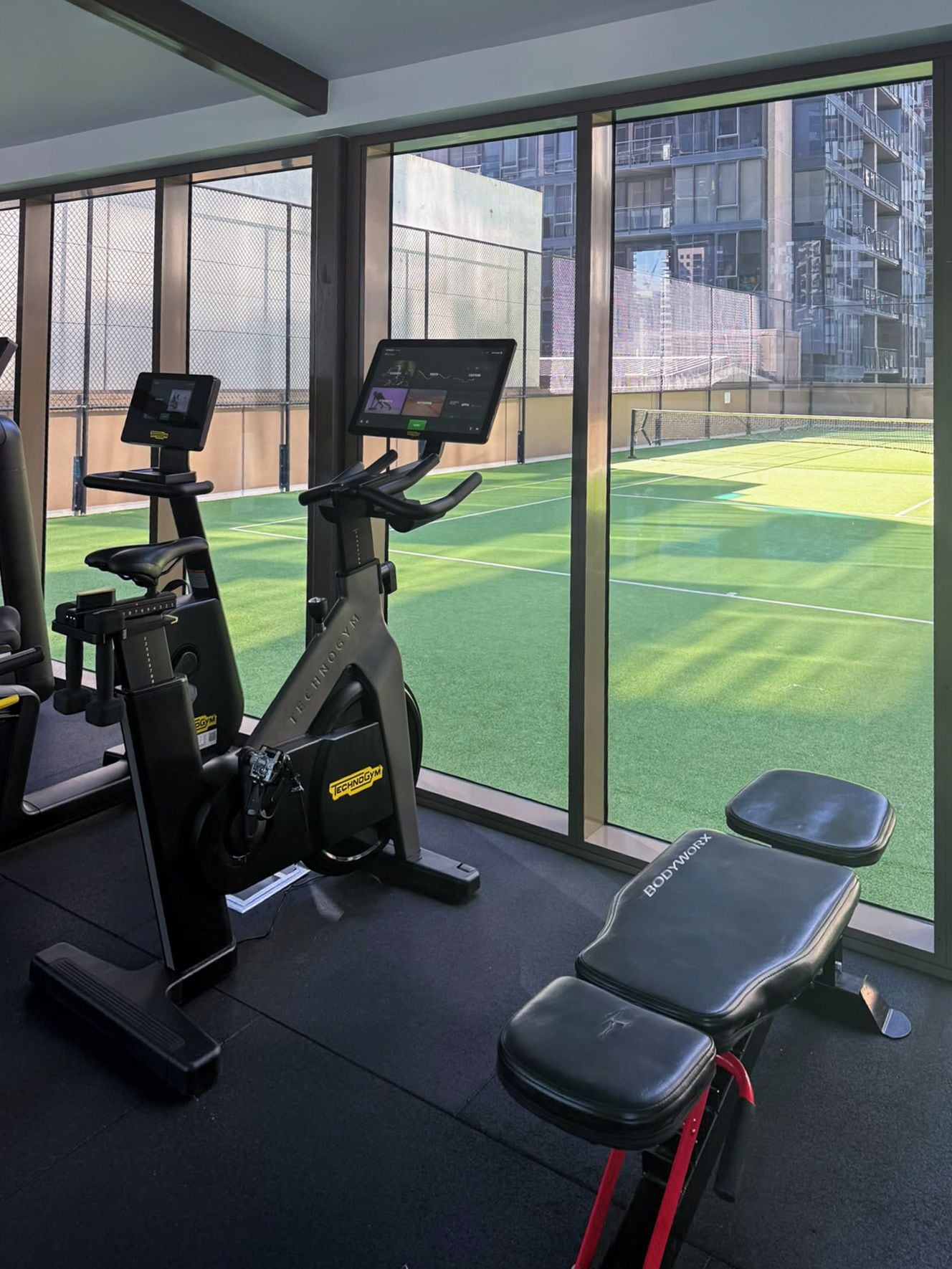 A gym with cardio machines and a bench, large windows with a view of a tennis court.