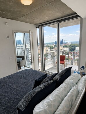 A modern bedroom with a large bed, a view of the city through floor-to-ceiling windows, and a balcony with a red chair.