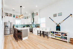 A modern kitchen with white cabinetry, a central island with bar stools, and a dining area with a white table and chairs. - Beachfront | 5BR | Sleeps 18 | Gulf Decks (Galveston)