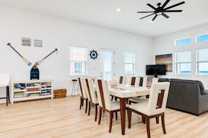 A modern living room with a dining area, wooden flooring, a television mounted on the wall, and a view of the outdoors through large windows. - Beachfront | 5BR | Sleeps 18 | Gulf Decks (Galveston)