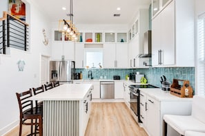 A modern kitchen with white cabinetry, a central island, and a dining area with wooden chairs. - Beachfront | 5BR | Sleeps 18 | Gulf Decks (Galveston)