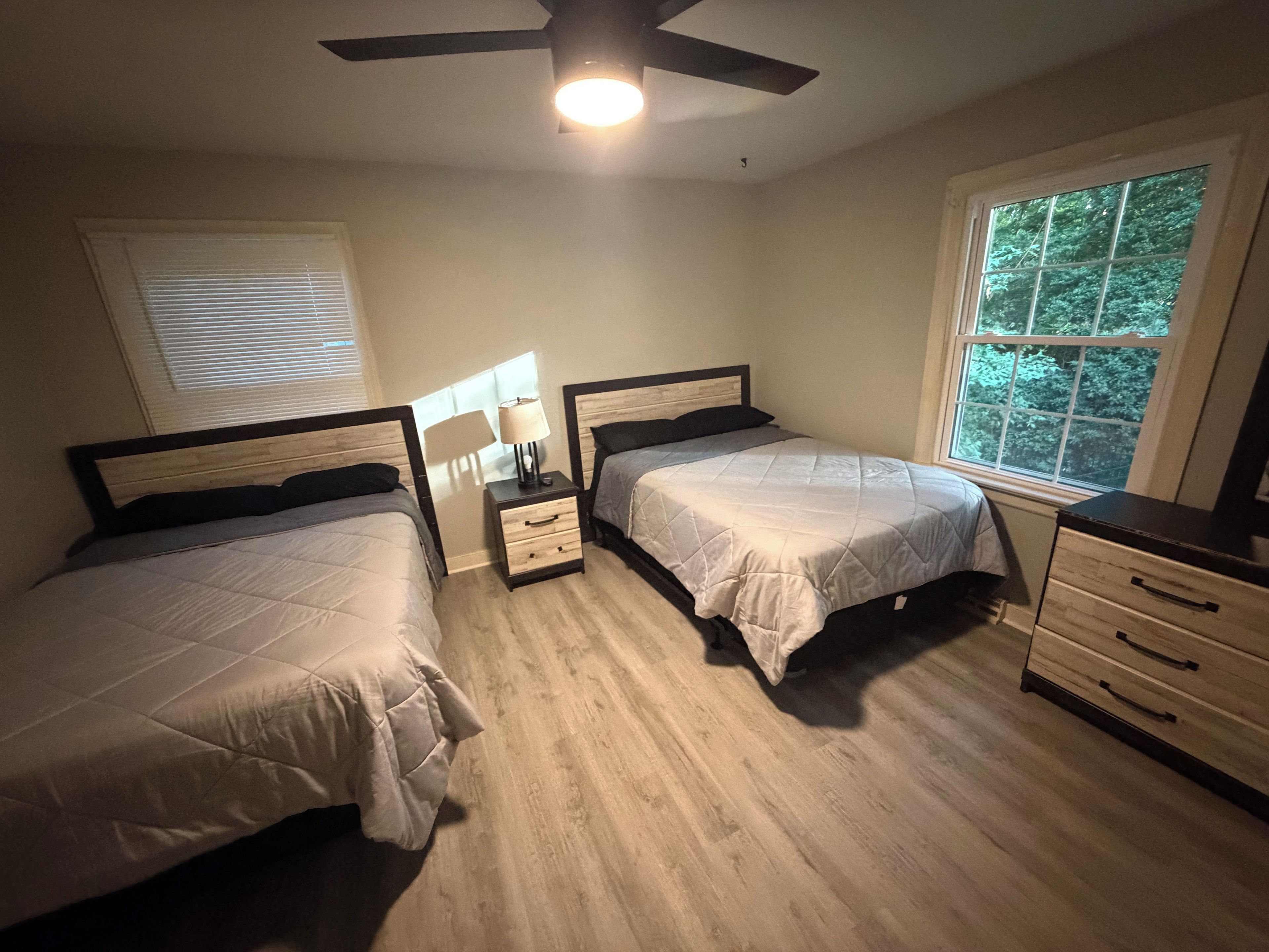 A bedroom with two beds, a nightstand with a lamp, a dresser, and a window with a view of trees.