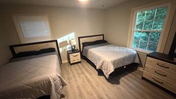 A bedroom with two beds, a nightstand with a lamp, a dresser, and a window with a view of trees.