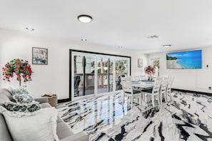 A modern dining area with a glass door leading to an outdoor balcony, white furniture, and a large TV on the wall. - Orange Blossom Cottage at White Sands/sleeps 8 steps to beach (Indian Rocks Beach)
