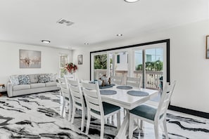 A dining area with a white table and chairs, a sofa with patterned cushions, and a large window with a view of greenery. - Orange Blossom Cottage at White Sands/sleeps 8 steps to beach (Indian Rocks Beach)