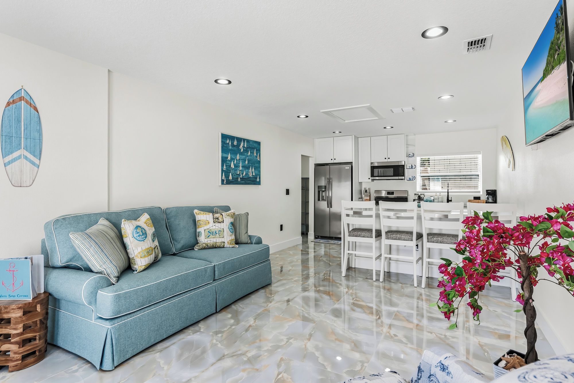 A modern living room with a blue sofa, a dining area with white chairs, a kitchen with a refrigerator, and a painting of sailboats on the wall.