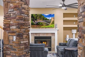 Een woonkamer met een open haard, een stenen muur, een televisie erboven en twee grijze banken.