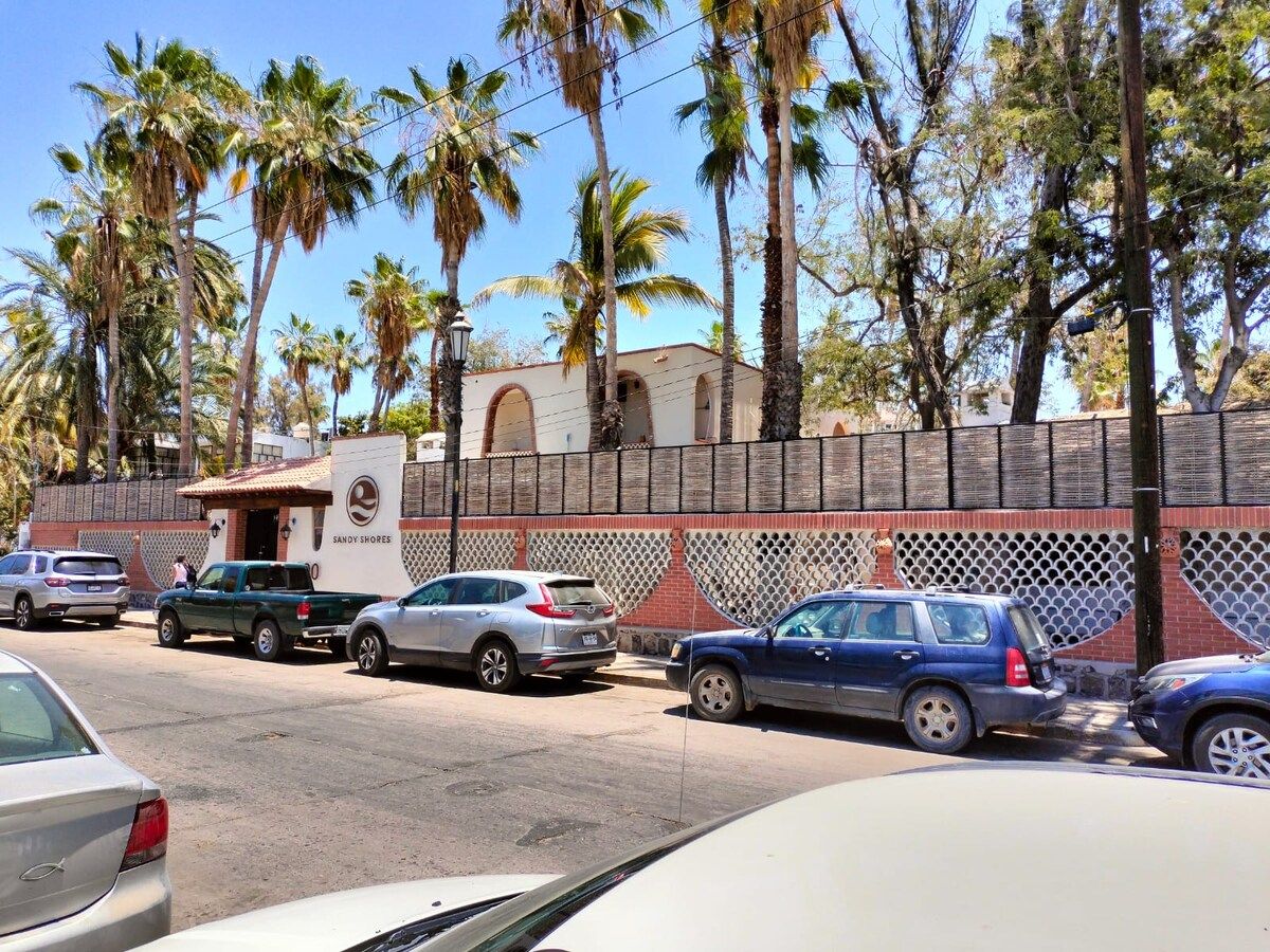 Un área de estacionamiento con varios autos, un edificio con un letrero que dice 'Sandy Shores' y una cerca con diseño de celosía.